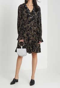 Maskáčové zavinovací šaty s volánkovým výstřihem a rukávy, černé nazouváky, drží malou bílou kabelku s kruhovým držadlem.