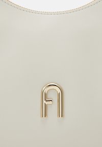 Borsa a mano in pelle beige con una texture liscia e un logo centrale in metallo dorato. Presenta un sottile bordo superiore cucito.