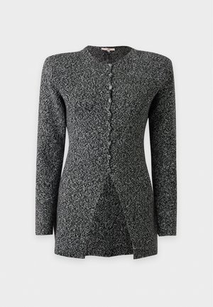 Un mannequin se tient devant un fond gris uni, vêtu d’un cardigan sombre texturé à boutons sur le devant, d’un pantalon noir et de chaussures à talons à brides.