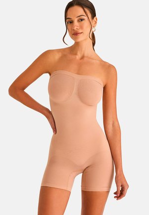 Femme portant un body gainant sans bretelles couleur nude avec un short, debout, une main sur la hanche, sur fond blanc.