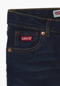 Tmavě modré džíny z denimu s měděnými nýty, žlutým prošíváním a červenou nášivkou loga Levi's na přední kapse. Hladký povrch.