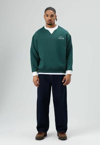 Groene trui met witte kraag en logo, gecombineerd met donkerblauwe jeans met wijde pijpen en bruine schoenen. Heeft een losse pasvorm en een textuurrijke stof.