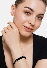 Bracciale nero con finitura lucida e design a catena; indossato su un polso, che mostra una texture liscia e un aspetto elegante.