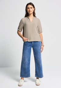 Beige, leichtes Blusenshirt mit V-Ausschnitt und gerafftem Saum; kombiniert mit weit geschnittenen blauen Jeans und weißen Sneakern.