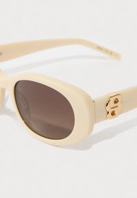 Κρέμα χρώματος οβάλ γυαλιά ηλίου με σκούρους φακούς και χρυσή διακοσμητική λεπτομέρεια "Β" στο βραχίονα του σκελετού.