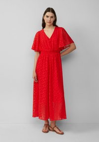 Vrouw in een rode midi-jurk met fonkelende mouwen en smock taille, gekleed in bruine platte sandalen tegen een effen lichtgrijze achtergrond.