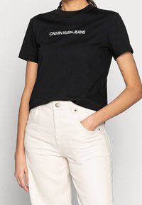T-shirt nera in cotone con la scritta "CALVIN KLEIN JEANS" in bianco. Design classico con scollo a girocollo e maniche corte. Abbinata a pantaloni beige chiaro.