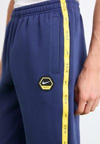 Sweatpants azul-marinho com cintura elástica, apresentando uma risca lateral amarela com o nome da marca impresso e um patch de logotipo hexagonal em preto e amarelo.