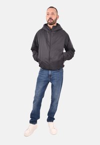 Giacca nera con zip frontale e cappuccio, polsini elasticizzati e texture liscia, abbinata a jeans in denim blu e sneakers bianche.