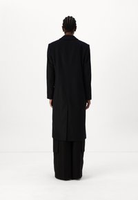 Lang sort frakke lavet af tekstureret stof, med knækket krave og en lige skæring, med minimal detaljerigdom, vist fra ryggen.
