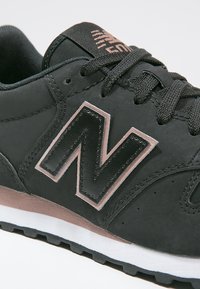 Baskets New Balance noires avec des accents gris et roses, semelle texturée et lacets noirs attachés en nœud.
