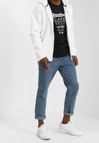 Valkoinen vetoketjuhuppari mustan graafisen T-paidan päällä, yhdistettynä vaaleansinihin kapeneviin farkkuihin ja valkoisiin tenneihin. Ominaisuuksiin kuuluu näkyviä logoja.