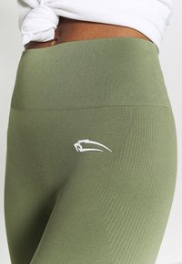 Leggings verde oliva hechos de una tela elástica que presentan una cinturilla acanalada y patrones geométricos tonales, con un logo blanco en el costado.