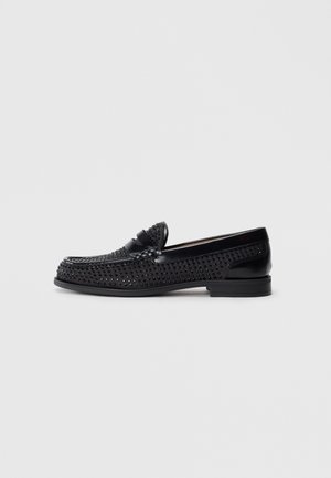 Mocassins en cuir noir avec un design perforé, un petit talon et une sangle décorative sur le devant. Fini lisse avec un bout arrondi.