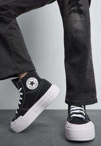 Sorte canvas høyhælte sneakers med hvit gummisåle og sømmer. Har Converse All Star-logo på siden. Brukt med svarte slitte jeans.