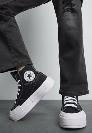 Baskets montantes en toile noire avec semelle en caoutchouc blanc et coutures. Présente le logo Converse All Star sur le côté. Portées avec un jeans noir déchiré.
