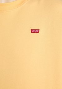 Camiseta de algodón amarilla con cuello redondo y un pequeño parche del logo rojo de Levi's en el lado izquierdo del pecho. Textura suave y diseño minimalista.