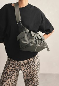 Luźna torba w oliwkowej zieleni z wieloma kieszeniami, wykonana z miękkiego materiału, z regulowanym paskiem i zapięciem na skręt. Noszona na czarnej bluzce i spodniach w cętki lamparta.