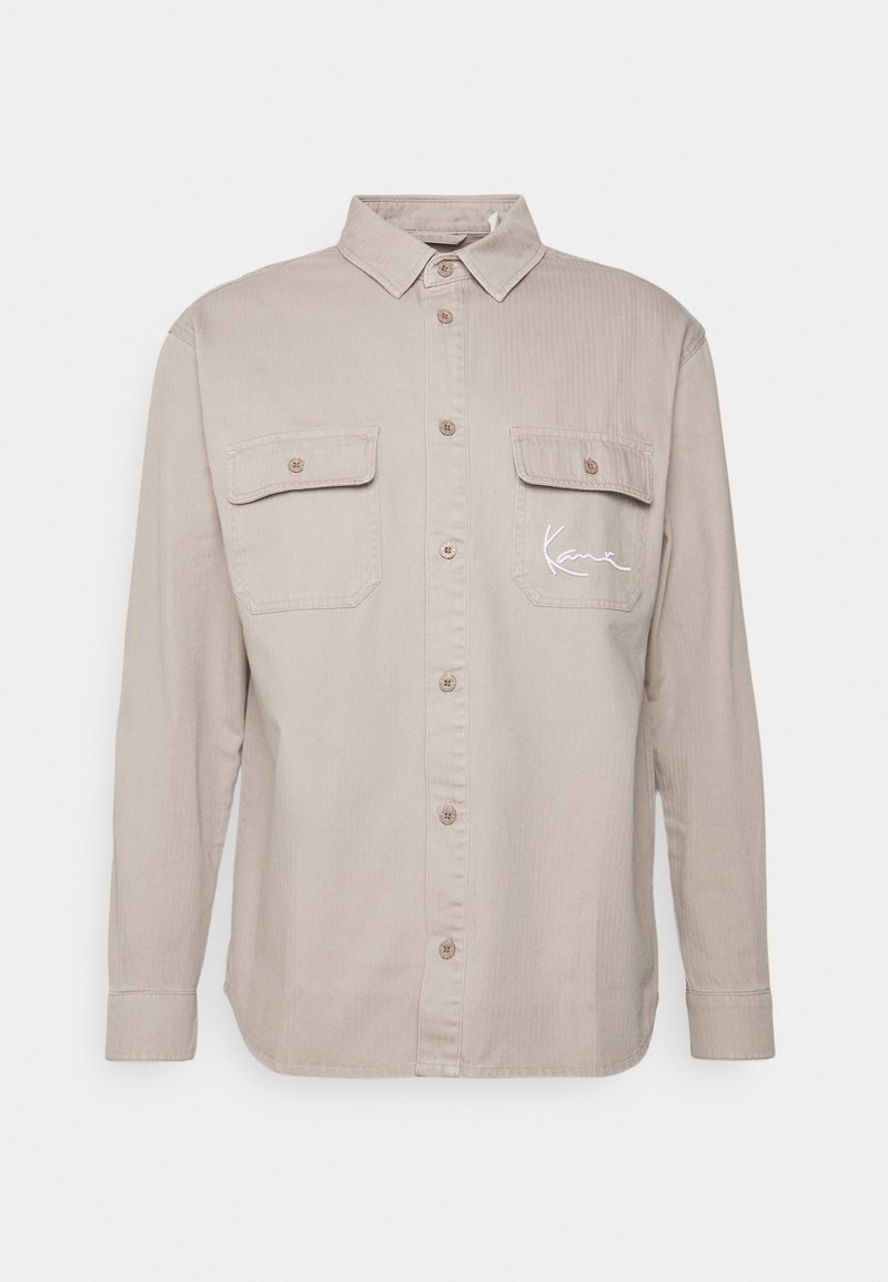 Beige långärmad skjorta med krage, två bröstfickor, knappstängning framtill och vit broderad logotyp på vänster ficka.