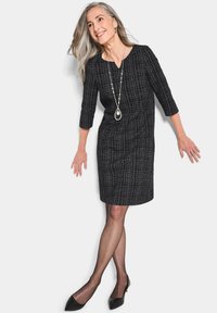 Robe à motif noir et blanc avec des manches trois-quarts, une longueur au genou et un décolleté en V, portée avec des escarpins noirs et un long collier.