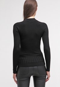 Pull noir côtelé à manches longues, coupe ajustée, col montant et motif en tissu texturé. Porté avec un pantalon noir ajusté.