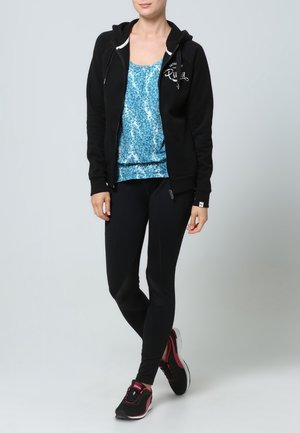 Donna in piedi che indossa una felpa nera con zip, un top senza maniche blu a fantasia, leggins neri e sneakers nere e rosa su uno sfondo grigio.