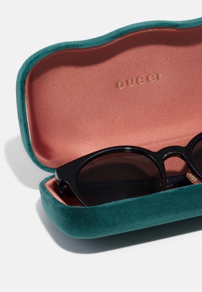 Czarne okulary przeciwsłoneczne spoczywające wewnątrz zielonego welwetowego etui z różową podszewką i złotymi literami "GUCCI" na pokrywie.