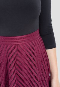 Falda burdeos con recortes en forma de chevron, forrada con una banda sólida, combinada con una blusa negra ajustada. El material parece tener textura.