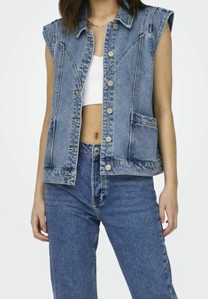 Vrouw draagt een mouwloos blauw denim vest met voorzakken en zilveren knopen, gecombineerd met hooggetailleerde blauwe spijkerbroek en wit cropped top.