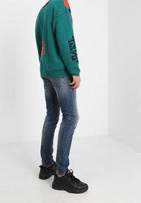 Teal stickad tröja med färgglada mönster, tillsammans med slim-fit blå jeans och svarta sneakers, som visar en avslappnad outfit bakifrån.