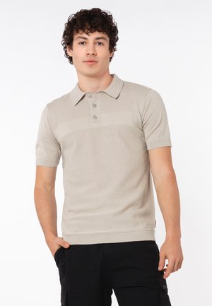Junger Mann mit lockigem Haar trägt ein beiges kurzärmeliges Poloshirt und schwarze Cargohosen, steht mit einer Hand in der Tasche vor weißem Hintergrund.