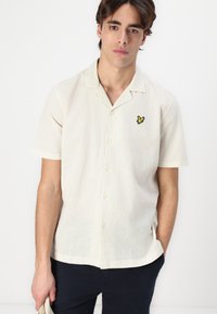Camicia a maniche corte di un leggero beige con una texture delicata, caratterizzata da una piccola farfalla gialla ricamata sul petto sinistro.
