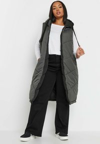 Lång, vadderad väst i mörkgrått med huva, dragkedjestängning och sidfickor. Buren över en vit topp och svarta byxor. Mjuk textur, quiltad design.