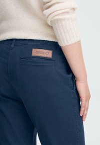 Mornarsko modri hlače z gladko teksturo, ki imajo zadnji žep in usnjen logotip z oznako "OXMO." Obleka je v kompletu s svetlo puloverjem.