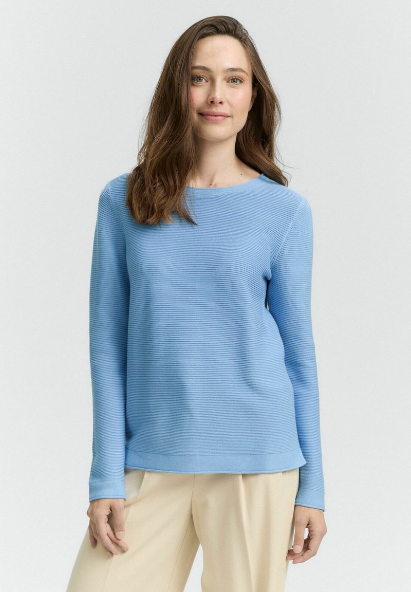 Pull bleu clair à manches longues avec texture côtelée, col rond et coupe décontractée, associé à un pantalon crème.