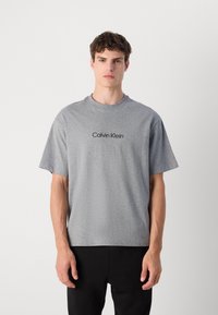 Grå t-skjorte med kort erm og rund hals, med en svart "Calvin Klein"-logo foran. Stoffet ser mykt og uformelt ut.