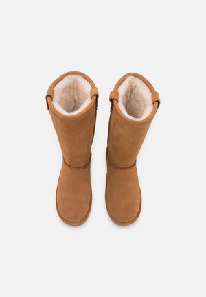 Bottes en suede marron jusqu'au genou avec une doublure douce de couleur crème, présentant un design simple et un détail de sangle à l'arrière pour un ajustement sécurisé.