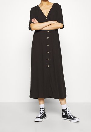 Personne portant une robe midi noire boutonnée à manches courtes, des chaussettes blanches et des baskets montantes noires, debout les bras croisés.