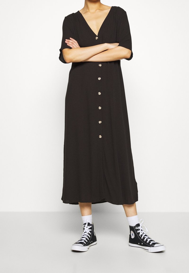 Personne portant une robe midi noire boutonnée à manches courtes, des chaussettes blanches et des baskets montantes noires, debout les bras croisés.