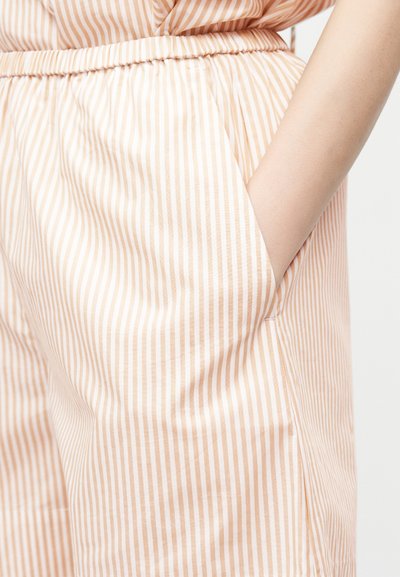 Χέρι στην τσέπη μπεζ και λευκού παντελονιού με κάθετες ρίγες, ελαστική μέση και ταιριαστό επάνω μέρος.