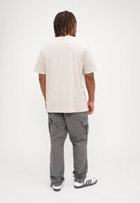 Beige katoenen T-shirt met korte mouwen, gecombineerd met grijze cargobroek met zakken, en witte sneakers met zwarte strepen.