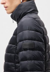 Melnā pūkainā jaka ar augstu apkakli, izšūtu dizainu un teksturētu audumu. Satur zīmola uzlīmi kreisajā pusē netālu no apakšmalas.