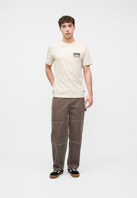 Νέος άνδρας που στέκεται φορώντας μπεζ μπλουζάκι με λογότυπο, άνετο καφέ παντελόνι με ραφές στα γόνατα και μαύρα αθλητικά παπούτσια με καουτσούκ σόλες.