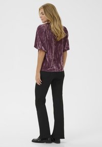 Kurzarmliges, gemustertes Oberteil aus tiefviolettem Samt, kombiniert mit schwarzen, schmalen Jeans und glänzenden schwarzen Schuhen. Hat einen lockeren Schnitt und sanft fallende Ärmel.