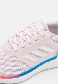 Jasnoróżowy but sportowy z siateczkową cholewką, białym logo z trzema paskami oraz podeszwą z gradientem od niebieskiego do różowego. Charakteryzuje się teksturowanymi sznurowadłami i lekkim designem.