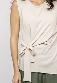 Haut sans manches beige en tissu texturé, avec un détail noué à la taille, un col rond et une coupe décontractée.