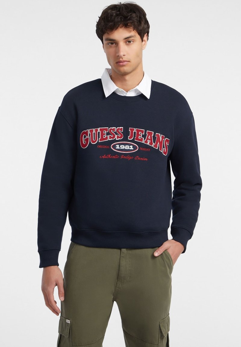 Sudadera azul marino con la inscripción "GUESS JEANS" en letras rojas y blancas. Cuello redondo y puños acanalados, llevada sobre una camisa blanca con cuello.