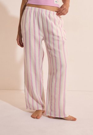 Persona que lleva pantalones sueltos y ligeros con rayas verticales en rosa pastel, verde y blanco, de pie descalza sobre un suelo claro.