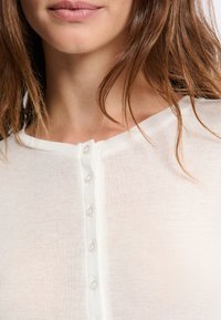 Haut blanc léger en maille avec un col arrondi et six petits boutons transparents sur le devant. Texture douce et aspect légèrement transparent.