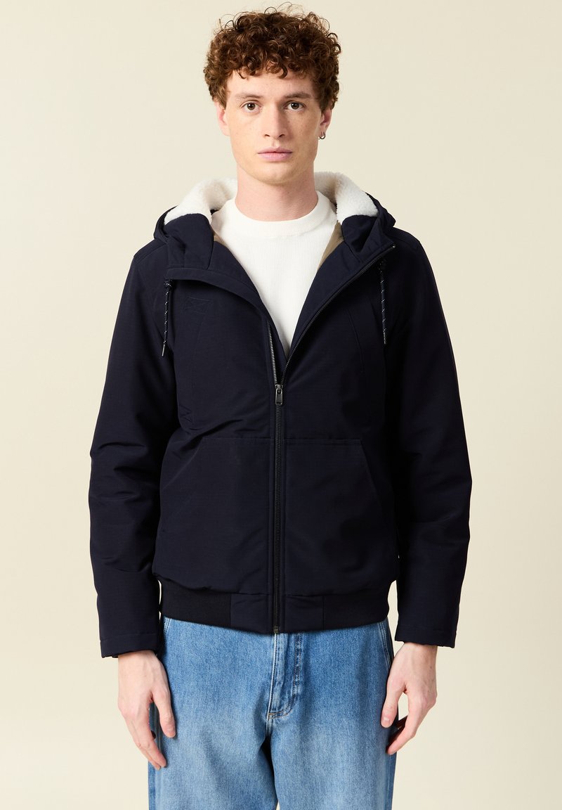 Giacca a vento blu navy con zip frontale, colletto foderato in pile e polsini elasticizzati, abbinata a jeans azzurri.